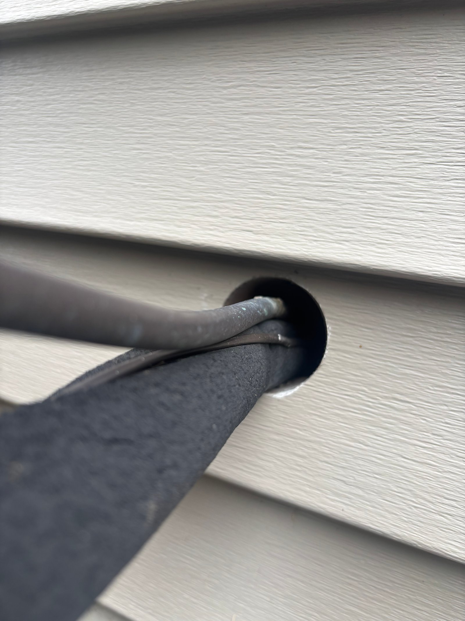 An open gap around the A/C refrigerant lines where they pass through vinyl siding, with bare space around the conduit