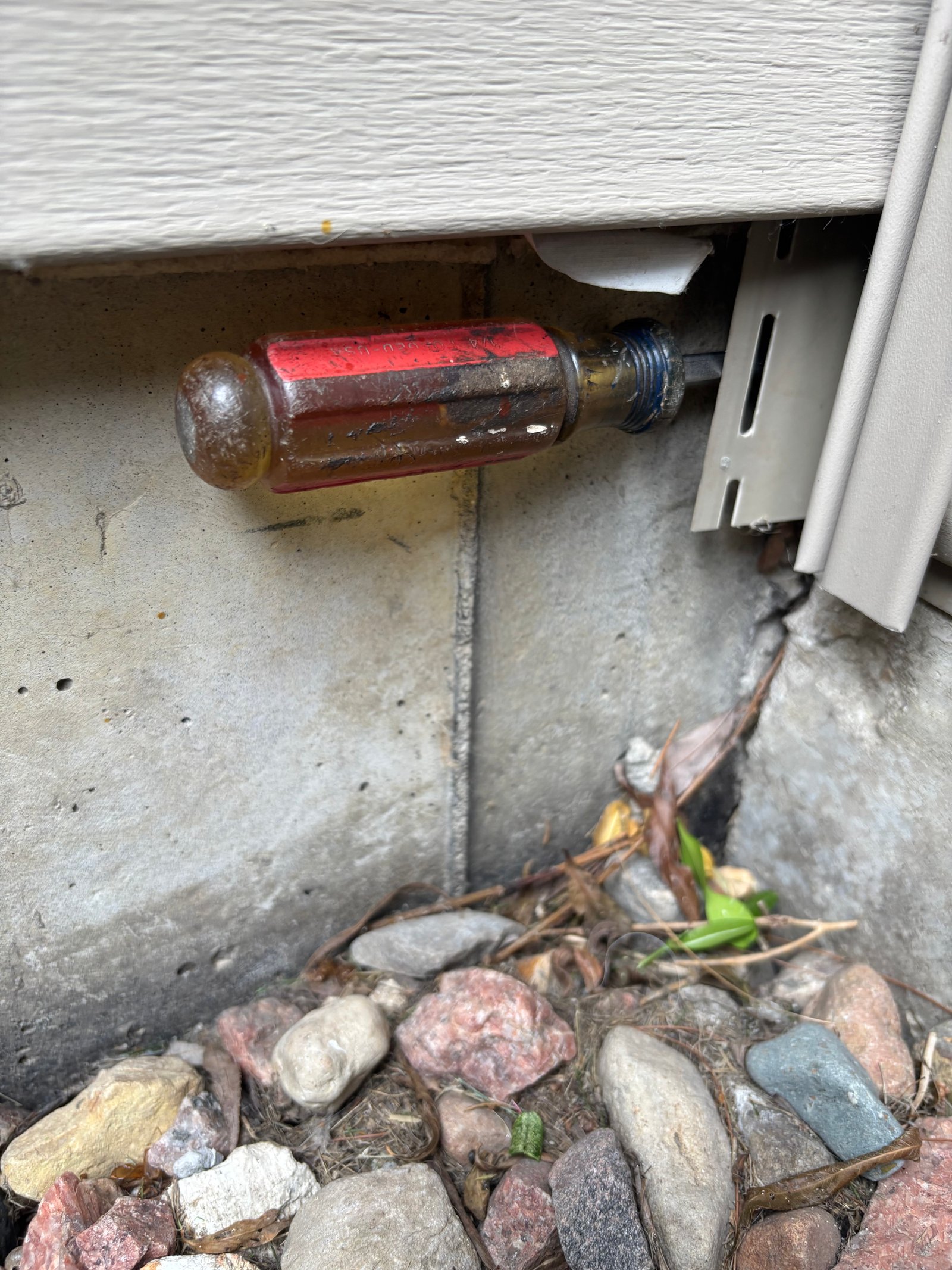 A screwdriver probing the gap where the bottom edge of vinyl siding meets a concrete stoop during an exterior inspection